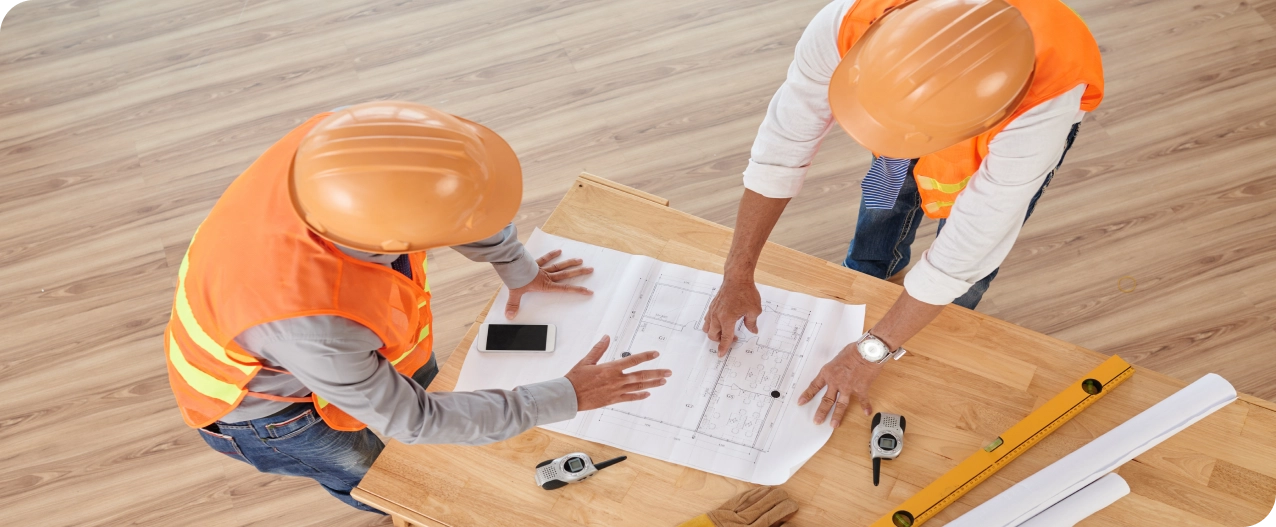 Des ouvriers en tenues de travail travaillent ensemble. Ils sont en train de discuter autour d'un plan de chantier posé sur une table. Chacun porte un gilet orange et un casque de sécurité.
